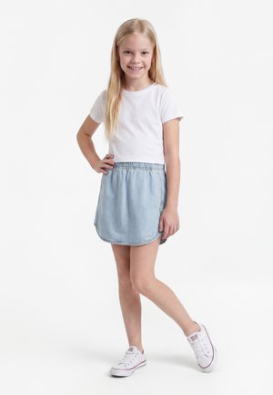 Młoda dziewczyna z długimi blond włosami, ubrana w białą koszulkę, jasnoniebieską spódnicę Nike i białe trampki, stoi z ręką na biodrze i uśmiecha się.