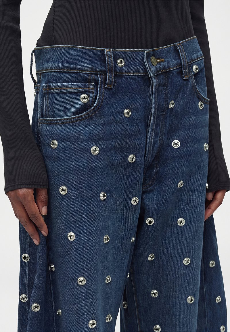 Jean en denim bleu foncé avec plusieurs œillets métalliques argentés couvrant le tissu, porté avec un haut noir à manches longues.