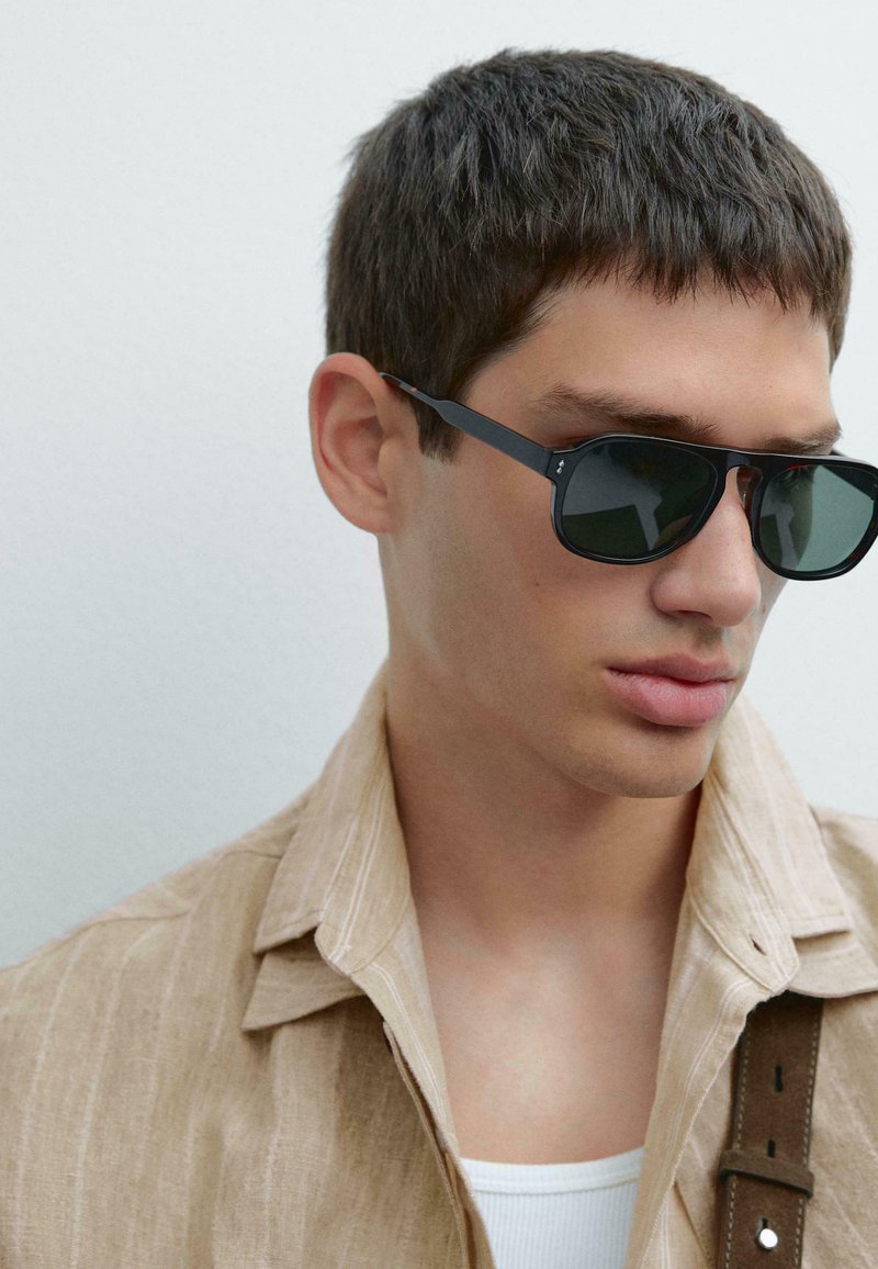 Černé sluneční brýle s geometrickým rámečkem, tmavé čočky, nošeny s světle béžovou lněnou košili s vertikálními pruhy a hnědým detailem na popruhu.