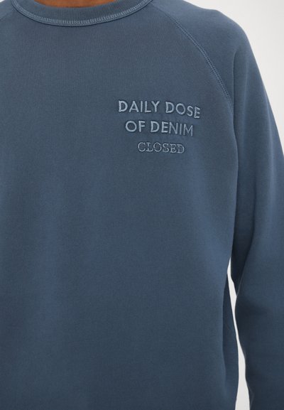 Pulover bleumarin cu mâneci raglan, având brodat textul "DOZA ZILNICĂ DE DENIM" și "ÎNCHIS" în fir de culoare albastru deschis. Textură netedă.