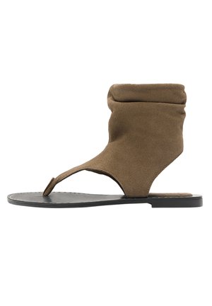 Brązowy zamszowy płaski sandał zawiązywany wokół kostki, z pętelką na palec i czarną podeszwą, zaprojektowany do noszenia na co dzień.
