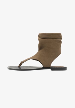 Brązowy zamszowy płaski sandał zawiązywany wokół kostki, z pętelką na palec i czarną podeszwą, zaprojektowany do noszenia na co dzień.