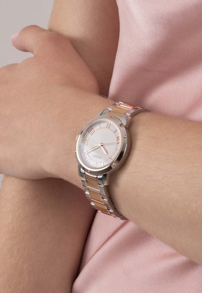 Strieborné a ružovo zlaté hodinky z nehrdzavejúcej ocele s bielym ciferníkom, rímskymi číslicami a dekoračnými akcentmi, zobrazené na svetlej pokožke zápästia.