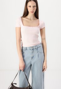 Vaaleanpunainen ribattupintainen toppi, jossa on lyhyet hihat ja neliön muotoinen pääntie, yhdistettynä korkeavyötäröisiin vaaleansinisiin väljälahkeisiin farkkuihin ja tumman käsilaukun kanssa.