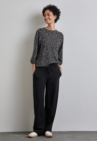 Schwarz-weißer gemusterter Langarmoberteil mit rundem Ausschnitt, kombiniert mit lockeren schwarzen Hosen und hellen Schuhen.