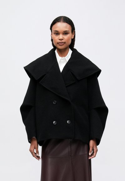 Donna che indossa un cappotto doppiopetto nero oversize con ampi rever sopra una camicia bianca e una gonna di pelle marrone scuro, in piedi contro uno sfondo neutro.
