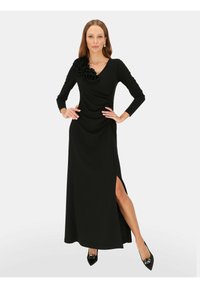 Czarna sukienka z długim rękawem, dekoltem w kształcie litery V, marszczonym designem, kwiatowym akcentem na ramieniu, rozcięciem po boku oraz gładką teksturą. Noszona z czarnymi szpilkami.