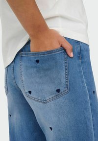 Jasnoniebieskie dżinsy z haftem w kształcie serca w granatowym kolorze na tylnych kieszeniach, z dopasowanym krojem i lekko przetartą teksturą.