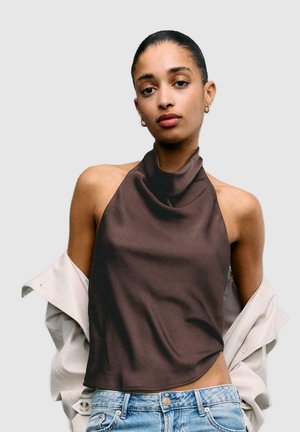 Femme aux cheveux lissés en arrière, portant un haut marron sans manches à col licou, une veste légère tombée sur les épaules et un jean en denim bleu, devant un fond uni.