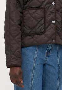 Veste matelassée marron avec un motif en losanges, comportant une grande poche et des boutons-pression. Les poignets ont une bordure marron contrastante.