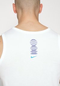 Man met een geschoren hoofd, gekleed in een witte mouwloze shirt met een blauw wereldbolpatroon en een lichtblauwe Nike swoosh op de bovenrug.