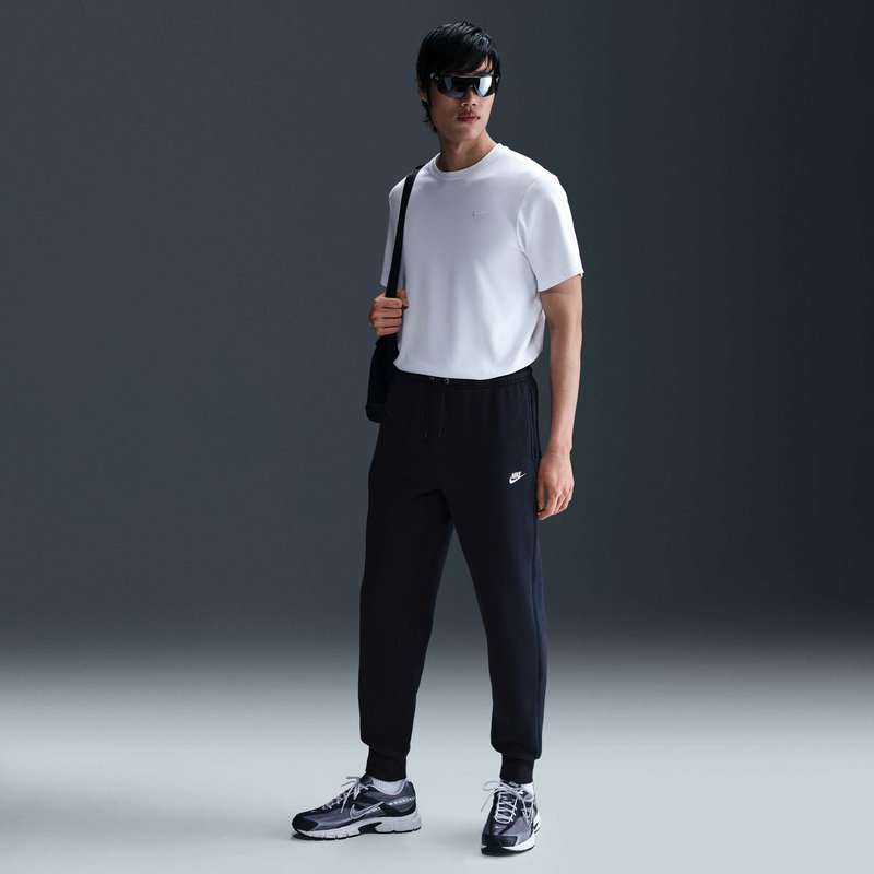 Uomo che indossa occhiali da sole, t-shirt bianca, pantaloni da tuta neri Nike, sneakers e una borsa a tracolla, in piedi contro uno sfondo scuro e uniforme.