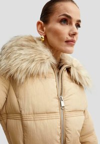 Beige puffjakke med en faux fur krave, quiltet struktur, lynlås i lukningen og sidesømme; har subtile syninger.