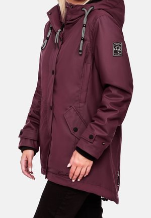 Femme portant un long imperméable violet avec des boutons noirs, des cordons à motifs sur la capuche, des poches avant et un écusson logo sur la manche gauche.