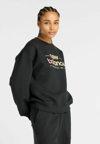 Sweatshirt noir avec col en côtes et coupe oversize. Présente le logo "new balance" en dégradé rose et jaune. Texture douce, design décontracté.
