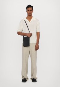 Sac en cuir noir porté en bandoulière avec une fermeture à rabat, de forme rectangulaire et une bandoulière réglable. Le modèle porte un polo en tricot de couleur claire et un pantalon beige.