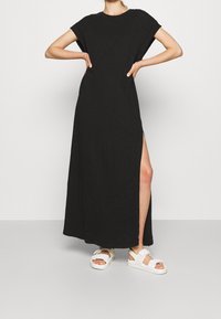 Černé maxi šaty s krátkým rukávem, boční rozparek na lemu, vyrobené z měkké tkaniny, doplněné bílými sandály na tenké pásky. Jednoduchý, volný střih.