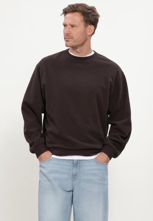 Uomo che indossa una felpa marrone scuro a maniche lunghe sopra una camicia bianca, con jeans azzurri, mani nelle tasche, guarda di lato.