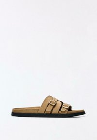 Hnědé semišové pantofle se dvěma nastavitelnými pásky a stříbrnými přezkami, s texturovanou černou gumovou podešví. Jednoduchý a plochý design.