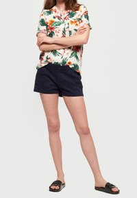 Kortärmad skjorta med knappar och tropisk blommig design i nyanser av grönt, orange och rosa; kombinerad med marinblå shorts och svarta sandaler.