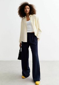 Crème oversized button-up shirt over een witte tanktop, gecombineerd met marineblauwe wijde broek en gele sneakers. Inclusief zwarte handtas.