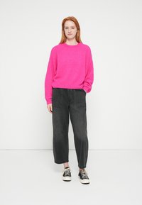 Fuksijas krāsas dzijas džemperis ar garām piedurknēm, kombinēts ar vaļīgām tumši pelēkām džinsa biksēm un melniem un baltiem auduma sporta apaviem.