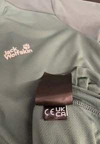 Veste Jack Wolfskin vert clair avec des étiquettes de conformité CE et UKCA visibles sur la couture intérieure près de la fermeture éclair.