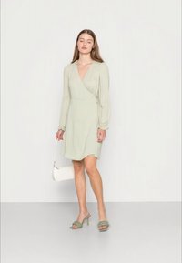 Vestido envolvente verde claro com mangas bufantes longas, textura suave e detalhe de nó na cintura. Combinado com sandálias com salto verdes e uma bolsa branca.