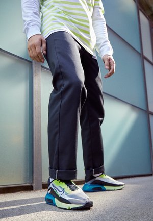 Sportieve sneakers met een grijze en zwarte bovenkant, neon groene accenten en een gestructureerde zool. Slim-fit zwarte broek met opgerolde pijpen.