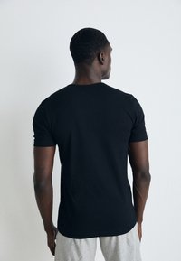 Schwarzes T-Shirt mit kurzen Ärmeln, aus glattem Baumwollstoff, körpernaher Schnitt, mit Rundkragen, von hinten gegen einen schlichten Hintergrund betrachtet.