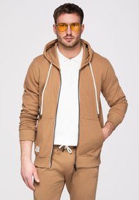 Homme portant un sweat à capuche beige zippé et un pantalon de survêtement, un polo blanc et des lunettes aviateur teintées de jaune, debout avec une main dans la poche.