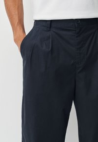 Tamsiai mėlynos spalvos kelnės iš lygaus audinio, su priekine klostė, sagų užsegimu ir šoninėmis kišenėmis papildomam patogumui.