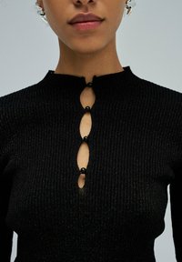 Maglione nero a coste con collo alto, dettaglio a chiave con perle nere e design aderente. La texture sembra morbida e leggermente luccicante.