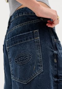 Close-up of hand resting on dark blue denim jeans with stitched patch reading "BDG" on back pocket.