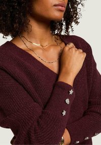 Maglione a maglia borgogna con scollo a V, caratterizzato da decorazioni di stelle e conchiglie argento sulle maniche. Indossato con collane di perline dorate e colorate.