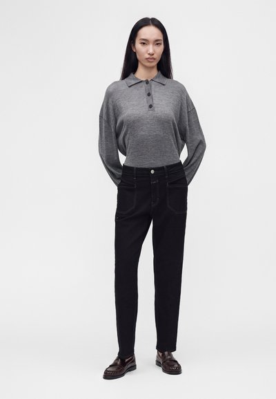 Femme debout portant un polo gris à manches longues, un jean taille haute noir et des mocassins marron foncé sur un fond blanc uni.