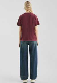 Vrouw met lang blond haar, gekleed in een bordeauxrode t-shirt met korte mouwen, losse blauwe spijkerbroek en witte sneakers, staande met haar rug naar de kijker op een effen achtergrond.