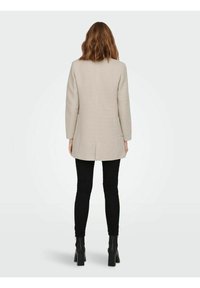 Beige dlouhý kabát se strukturovaným střihem, rovnou siluetou a detailem rozparku na zadní straně, v kombinaci s černými slim-fit kalhotami a kotníkovými botami.