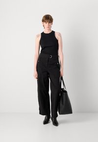 Giovane in top senza maniche nero, pantaloni neri a vita alta e larghi, stivali neri alla caviglia, che tiene una grande borsa nera, in piedi contro una parete bianca.