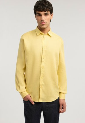 Junger Mann mit kurzen dunklen Haaren, der ein langärmliges gelbes Hemd und dunkle Hosen trägt und mit einer Hand in der Tasche steht.