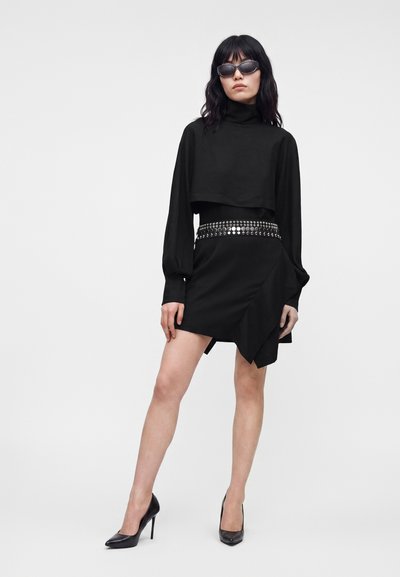 Femme portant un crop top noir à manches longues, une mini-jupe noire avec ceinture cloutée, des talons hauts noirs et des lunettes de soleil foncées sur un fond uni.