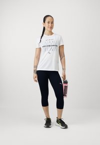 T-shirt blanc à manches courtes avec texte noir, legging cropped marine, chaussures noires avec accents néons, et une bouteille d'eau rose étiquetée "PUMA."