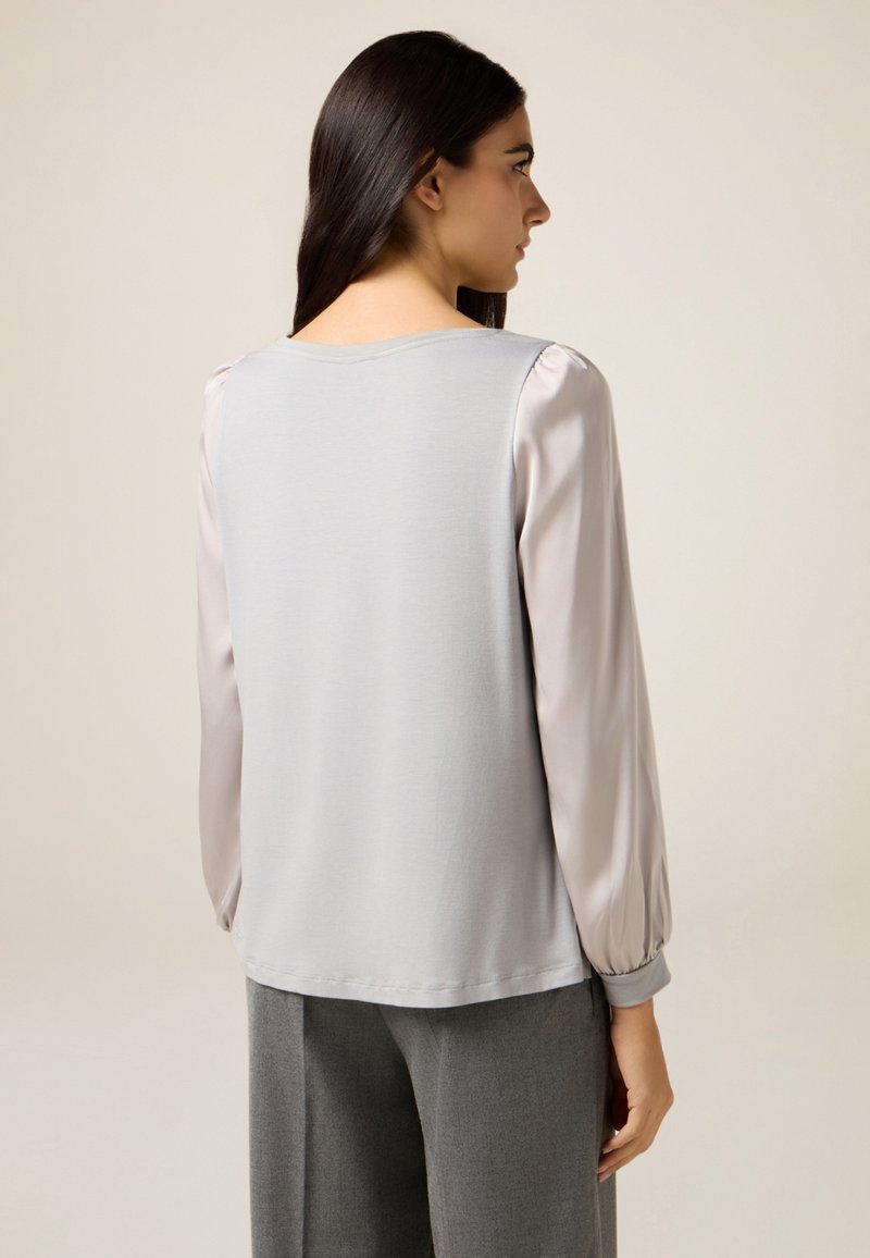 Top grigio con maniche lunghe e trasparenti, scollo arrotondato e corpino dalla texture liscia. La vista posteriore mostra cuciture sottili e una vestibilità comoda.