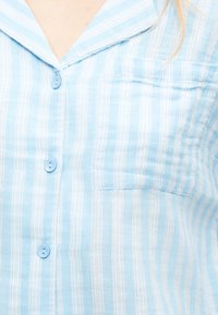 Chemise boutonnée à rayures verticales bleu clair et blanches, avec col et poche poitrine, portée par une personne.