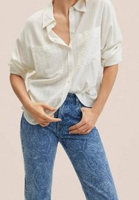 Camisa blanca de botón con textura y mangas arremangadas, combinada con jeans azules de patrón que presentan un diseño floral. Detalle de bolsillo frontal minimalista.