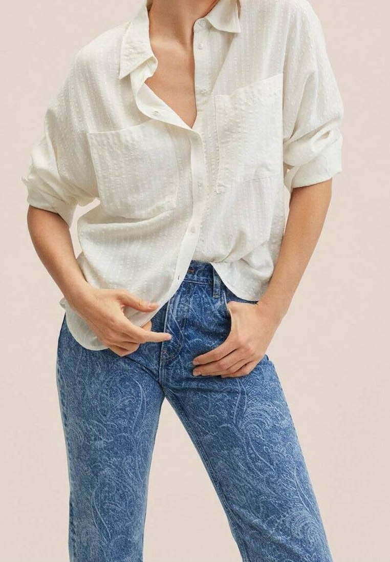 Camisa blanca de botón con textura y mangas arremangadas, combinada con jeans azules de patrón que presentan un diseño floral. Detalle de bolsillo frontal minimalista.