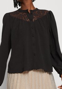 Blusa nera con un pannello in pizzo sulle spalle, con scollo rotondo, maniche lunghe e chiusura con bottoni sul davanti.