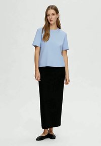 Ženska stoji oblečena v svetlo modro majico s kratkimi rokavi, dolgem črnem krilu in črnih balerinkah na enobarvni svetli podlagi.