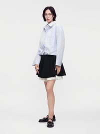 Femme portant une chemise oversize rayée bleu clair, une jupe plissée noire sur une jupe blanche superposée, et des chaussures Mary Jane noires, debout sur fond blanc.