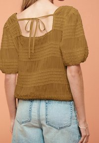 Olivengrøn bluse med teksturerede folder, korte pufærmer og en firkantet åbning bagpå, bundet med et bånd. Sammen med lyseblå denimshorts.
